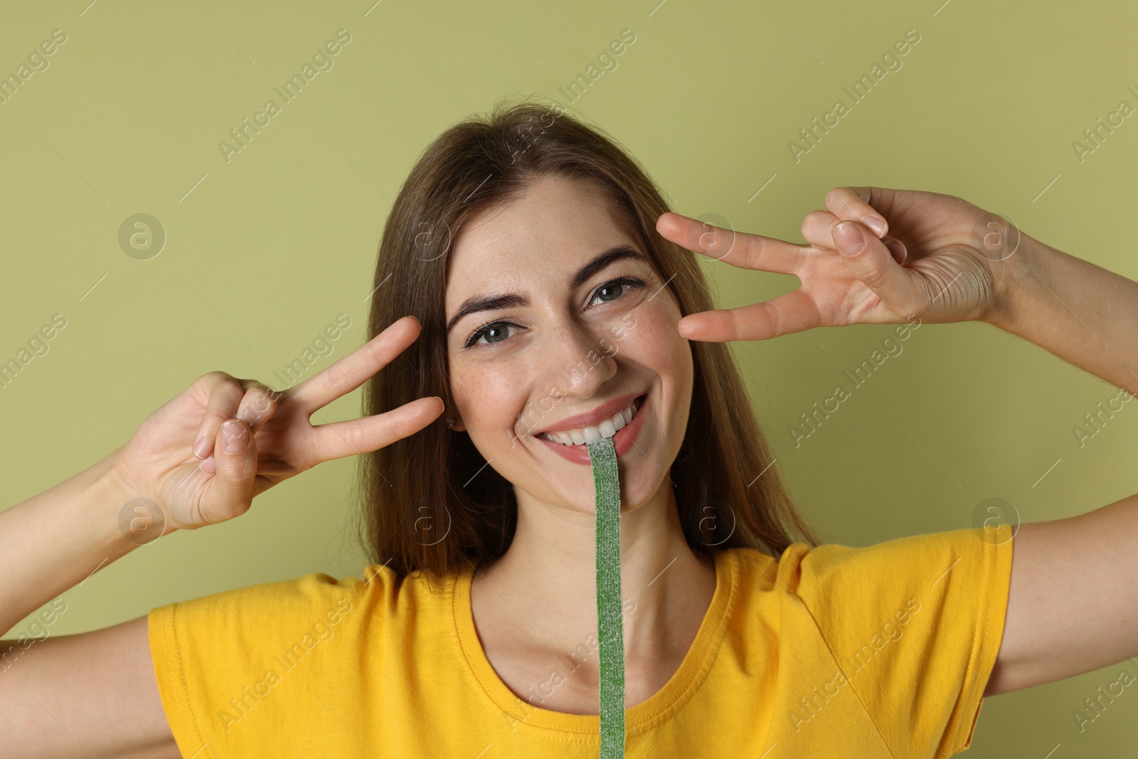 Photo of Happy young woman eating tasty gummy candy while showing v-sign on olive background