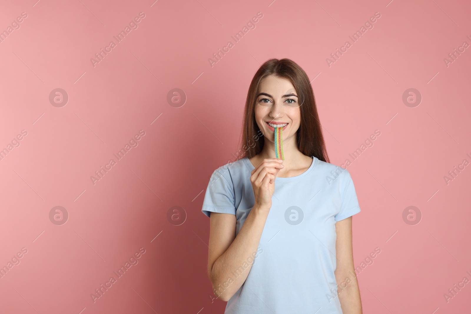 Photo of Young woman eating tasty rainbow sour belt on pink background, space for text