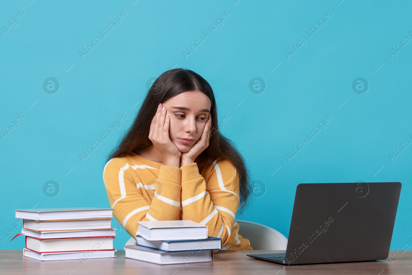 Photo of Young student with books and laptop having stress before exam at table against light blue background