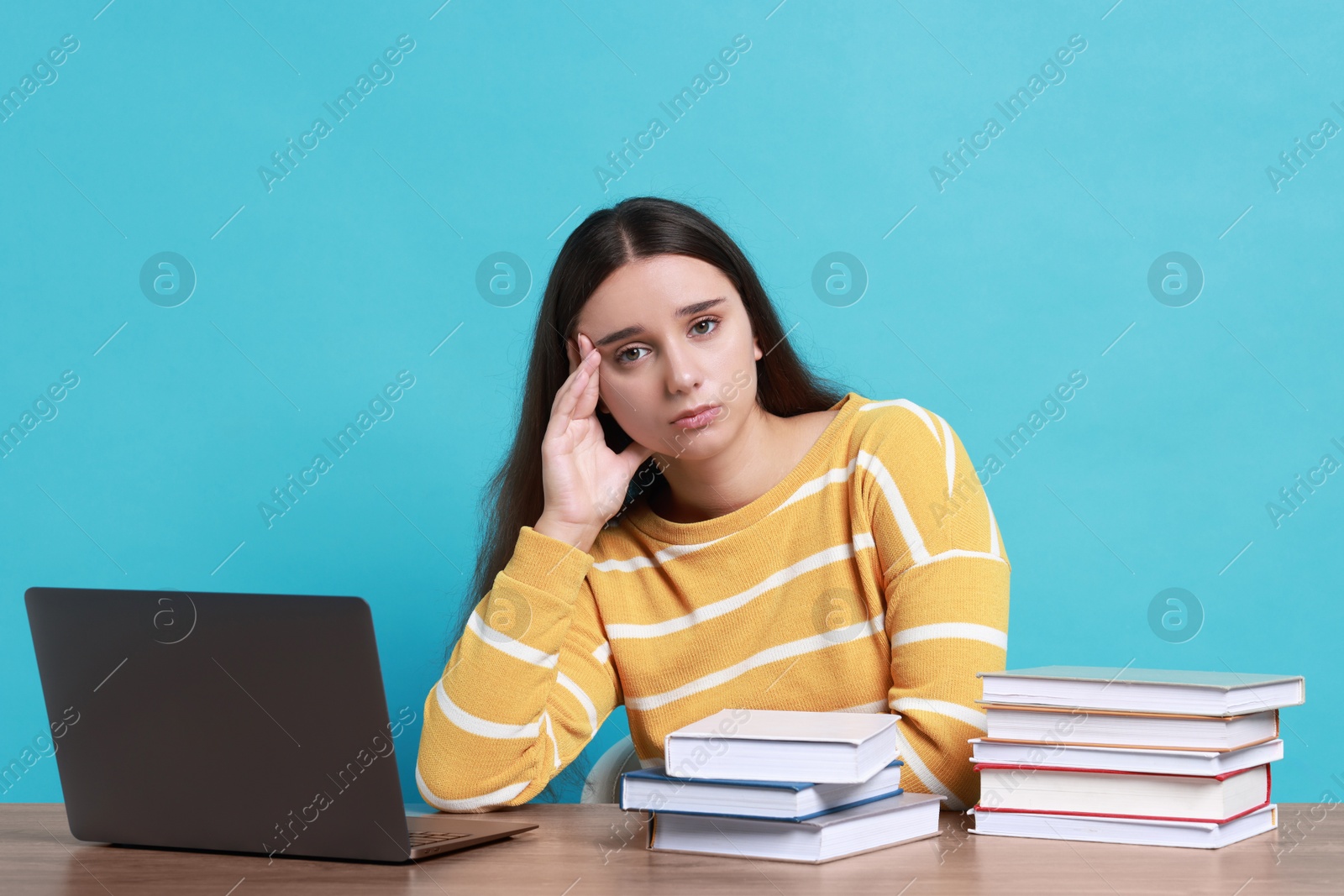 Photo of Young student with books and laptop having stress before exam at table against light blue background