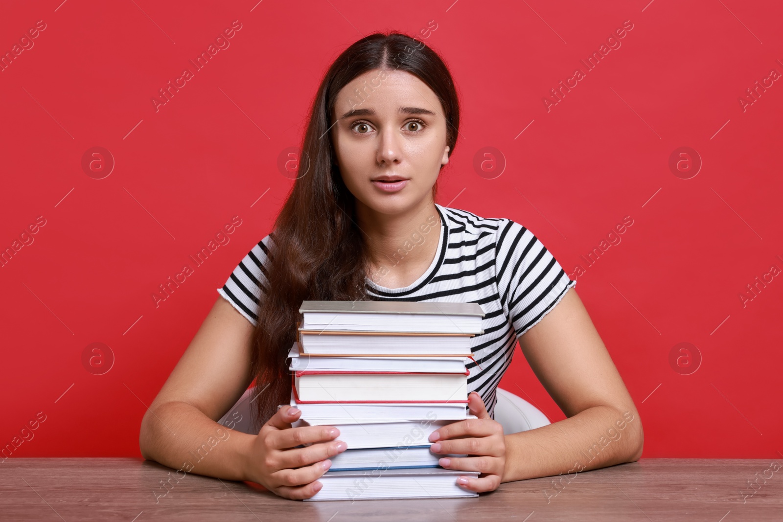 Photo of Young student with stack of books having stress before exam at table against red background