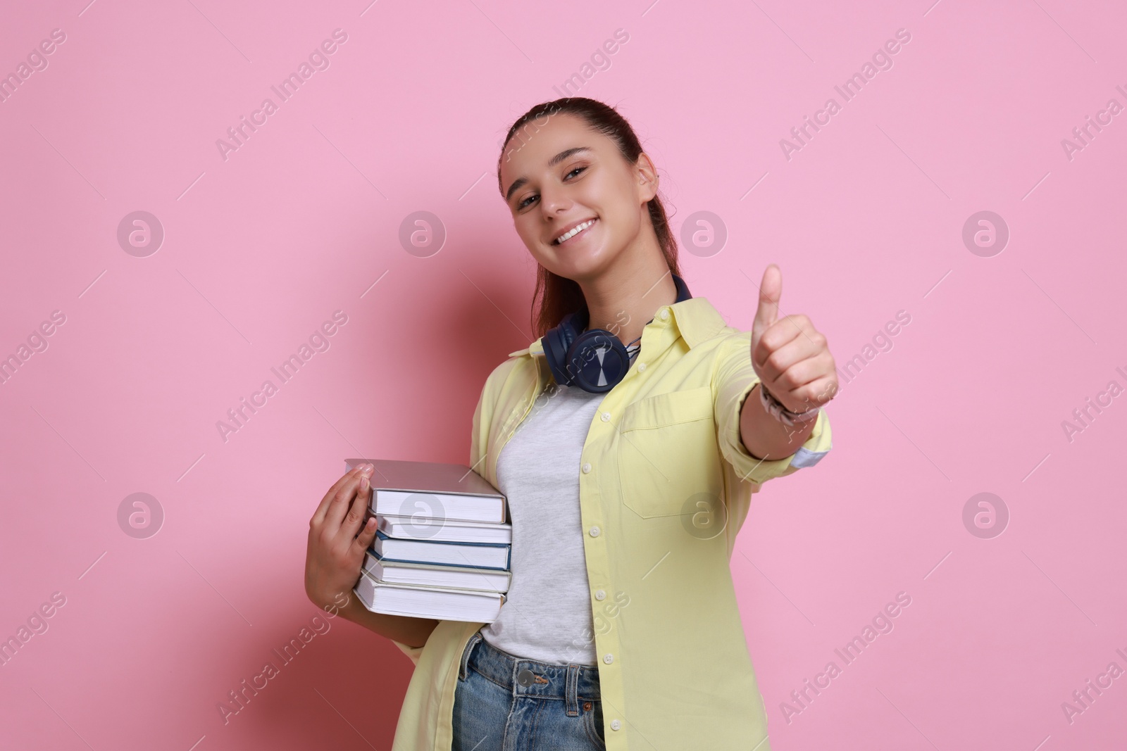 Photo of Happy young student with stack of books showing thumbs up on pink background