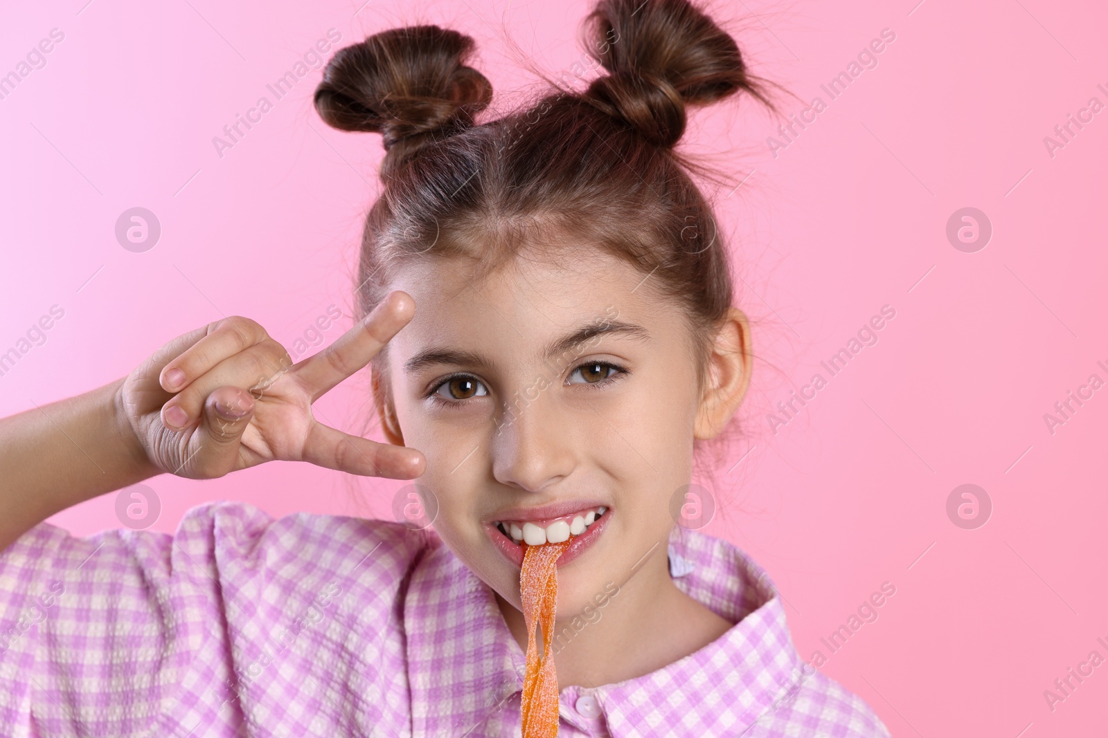 Photo of Happy girl eating tasty gummy candy and showing peace sign on pink background