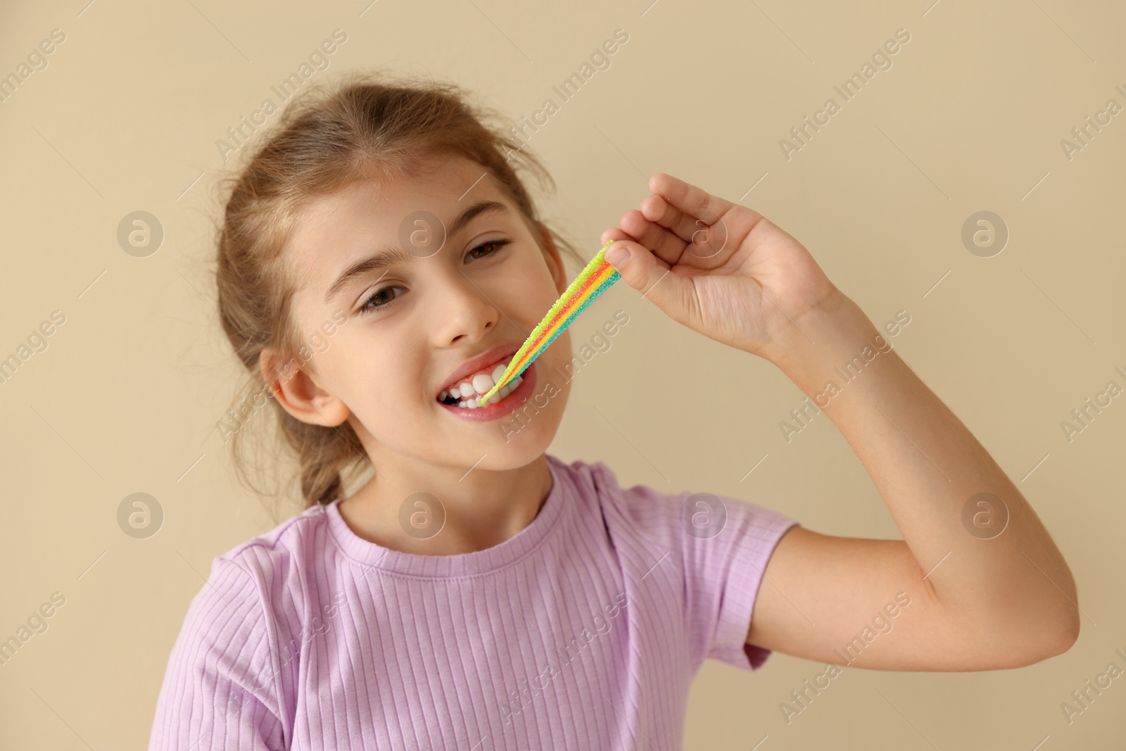 Photo of Little girl eating tasty rainbow sour belt on beige background