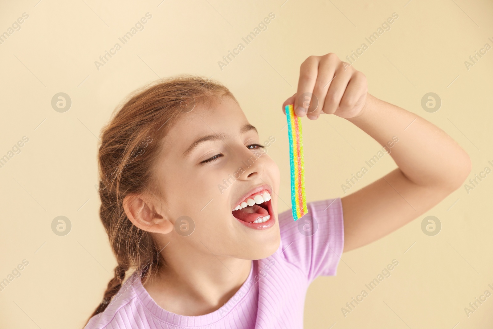 Photo of Little girl eating tasty rainbow sour belt on beige background