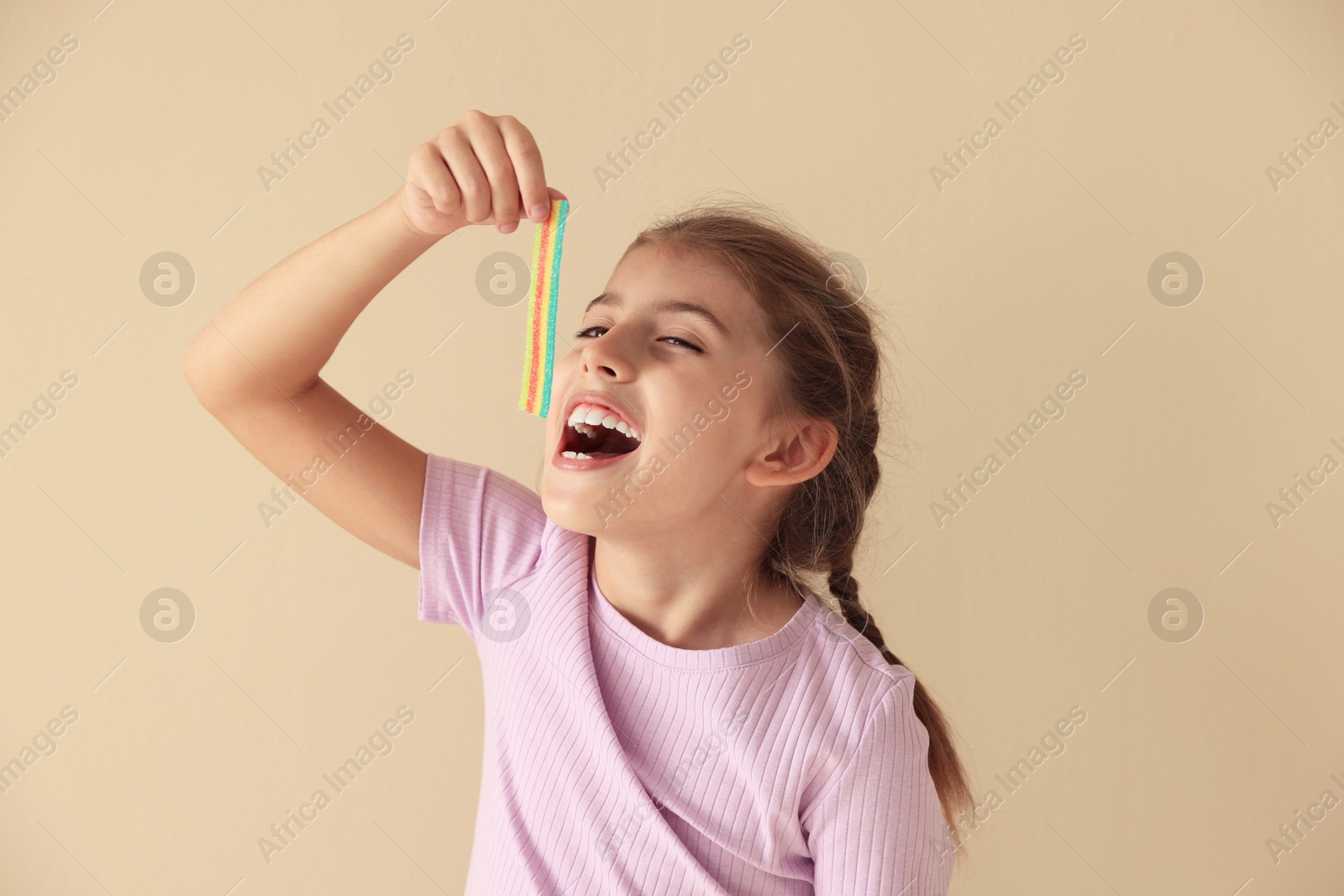 Photo of Little girl eating tasty rainbow sour belt on beige background