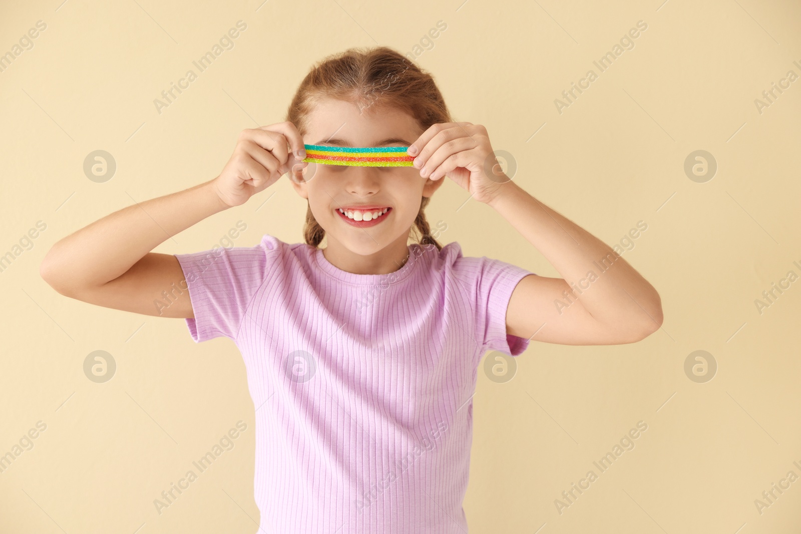 Photo of Happy girl covering eyes with tasty rainbow sour belt on beige background