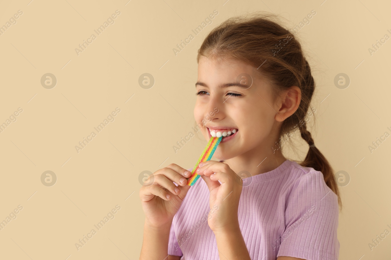 Photo of Happy girl eating tasty rainbow sour belt on beige background. Space for text