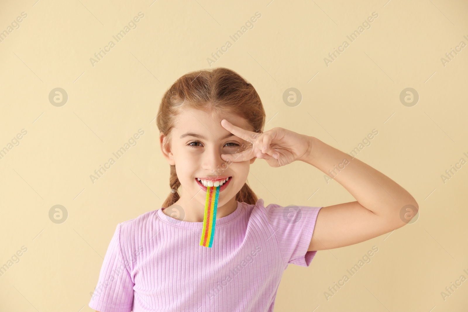 Photo of Happy girl eating tasty rainbow sour belt and showing peace sign on beige background