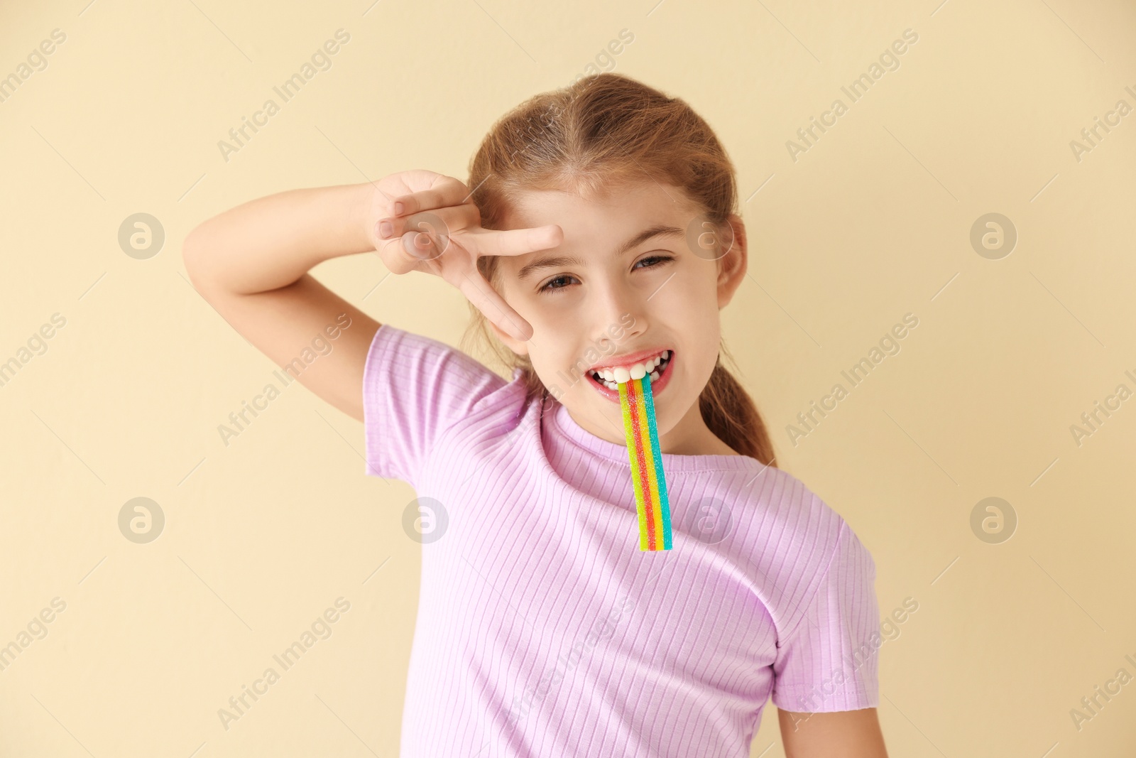 Photo of Happy girl eating tasty rainbow sour belt and showing peace sign on beige background