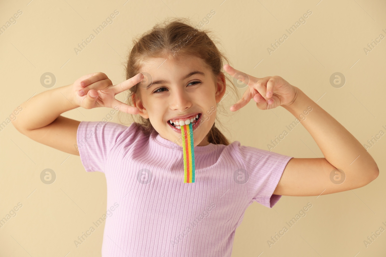 Photo of Happy girl eating tasty rainbow sour belt and showing peace signs on beige background