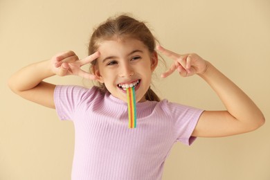 Photo of Happy girl eating tasty rainbow sour belt and showing peace signs on beige background