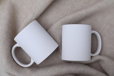 Photo of Blank white ceramic mugs on beige fabric, top view. Mockup for design