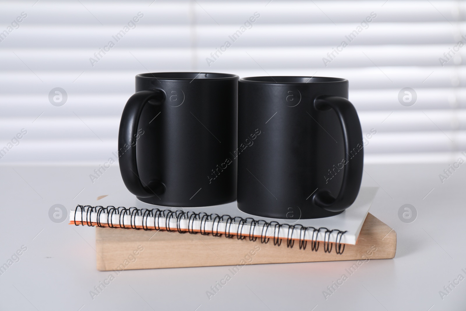 Photo of Blank black ceramic mugs and notebooks on white table. Mockup for design