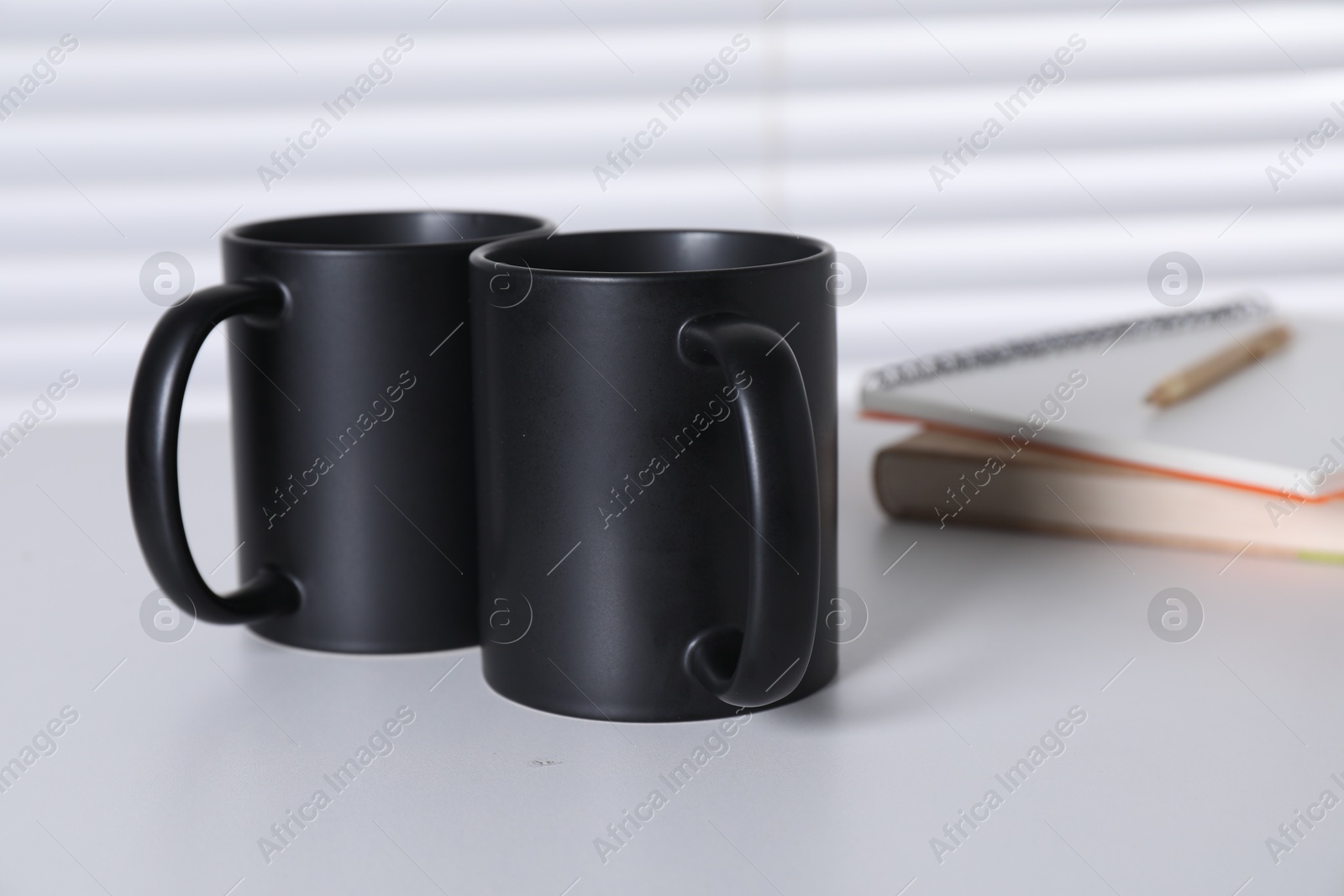 Photo of Blank black ceramic mugs and notebooks on white table. Mockup for design