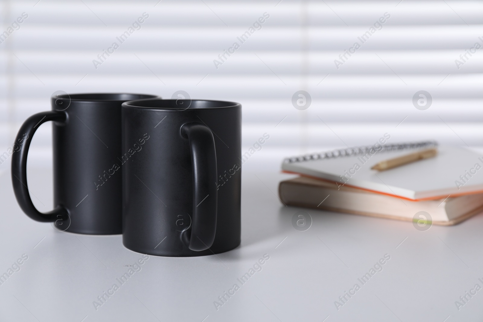 Photo of Blank black ceramic mugs and notebooks on white table. Mockup for design