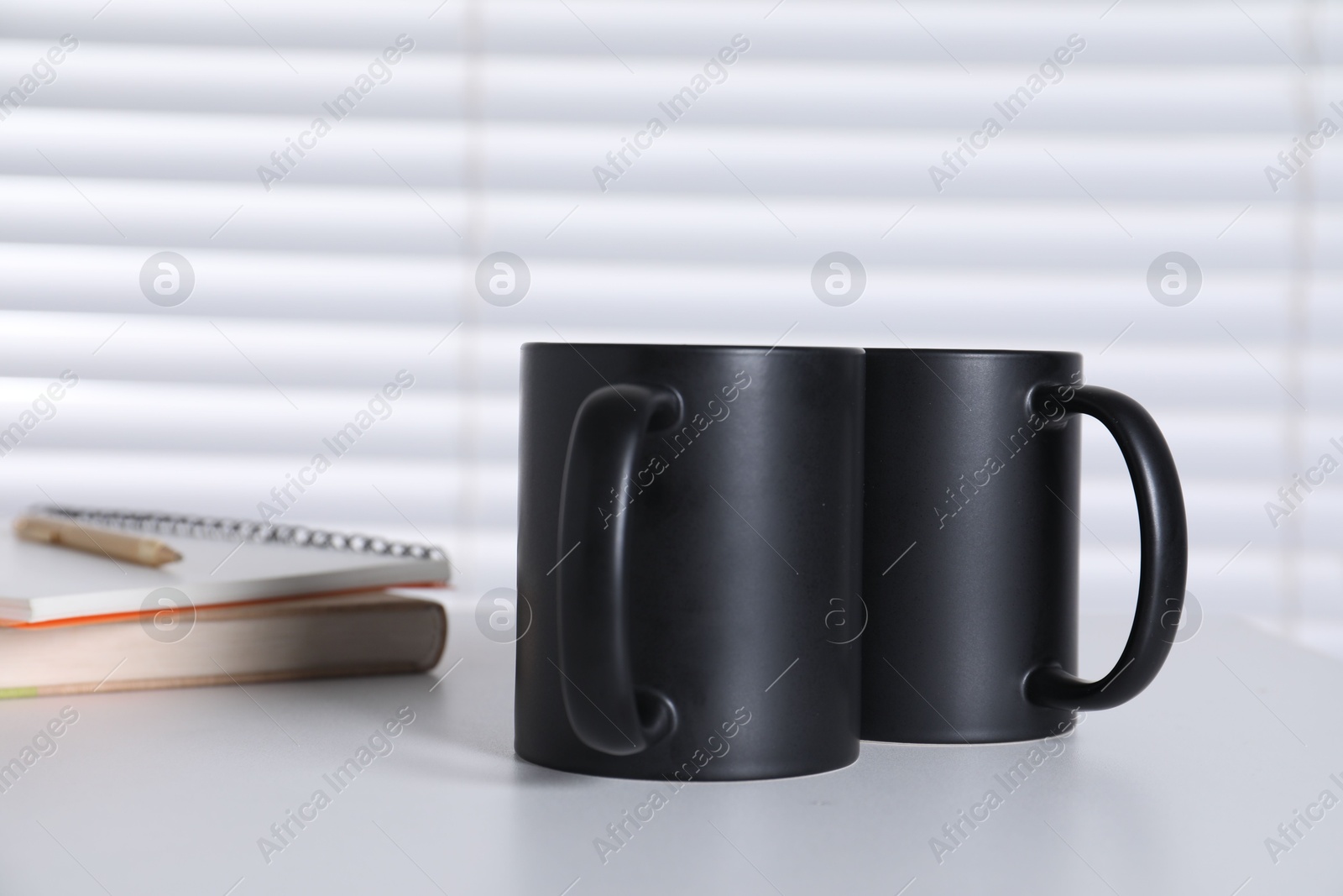 Photo of Blank black ceramic mugs and notebooks on white table. Mockup for design