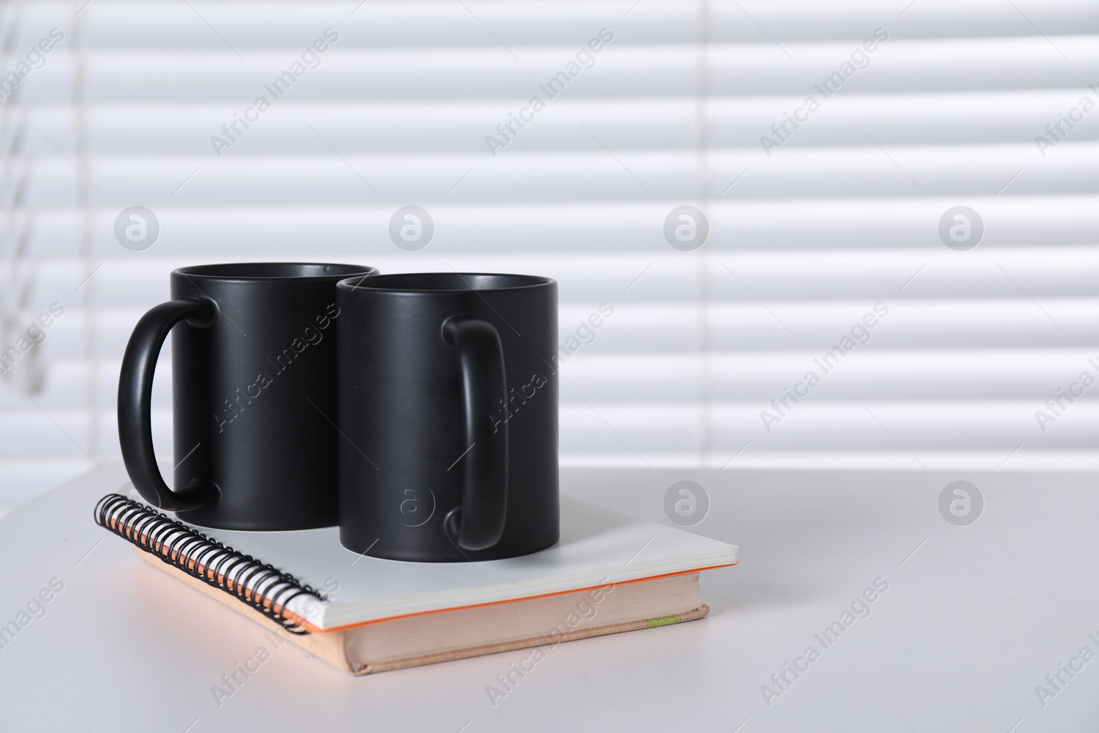 Photo of Blank black ceramic mugs and notebooks on white table. Mockup for design