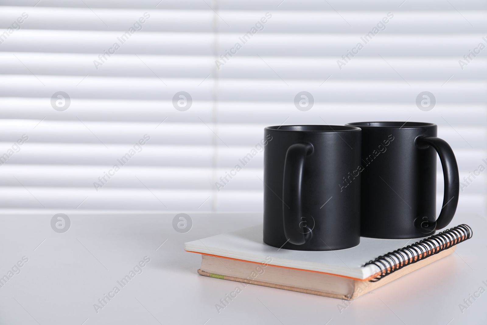 Photo of Blank black ceramic mugs and notebooks on white table. Mockup for design