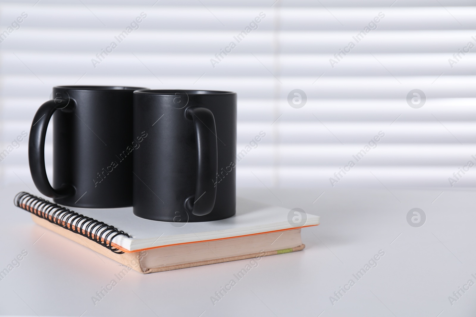 Photo of Blank black ceramic mugs and notebooks on white table. Mockup for design