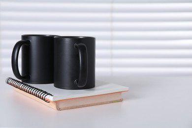 Photo of Blank black ceramic mugs and notebooks on white table. Mockup for design