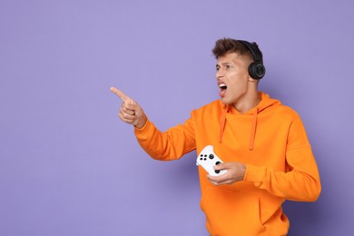 Emotional young man in headphones with controller on violet background, space for text