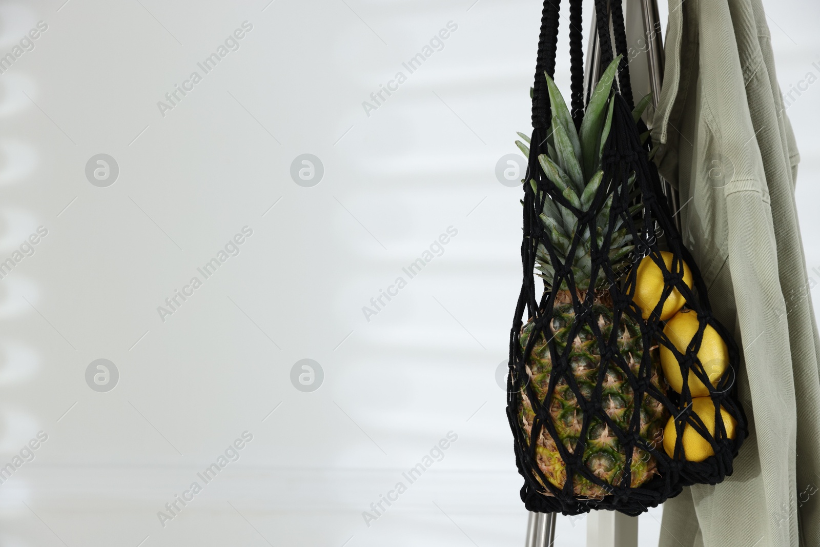 Photo of Macrame bag with fruits and jacket on clothing rack near white wall. Space for text