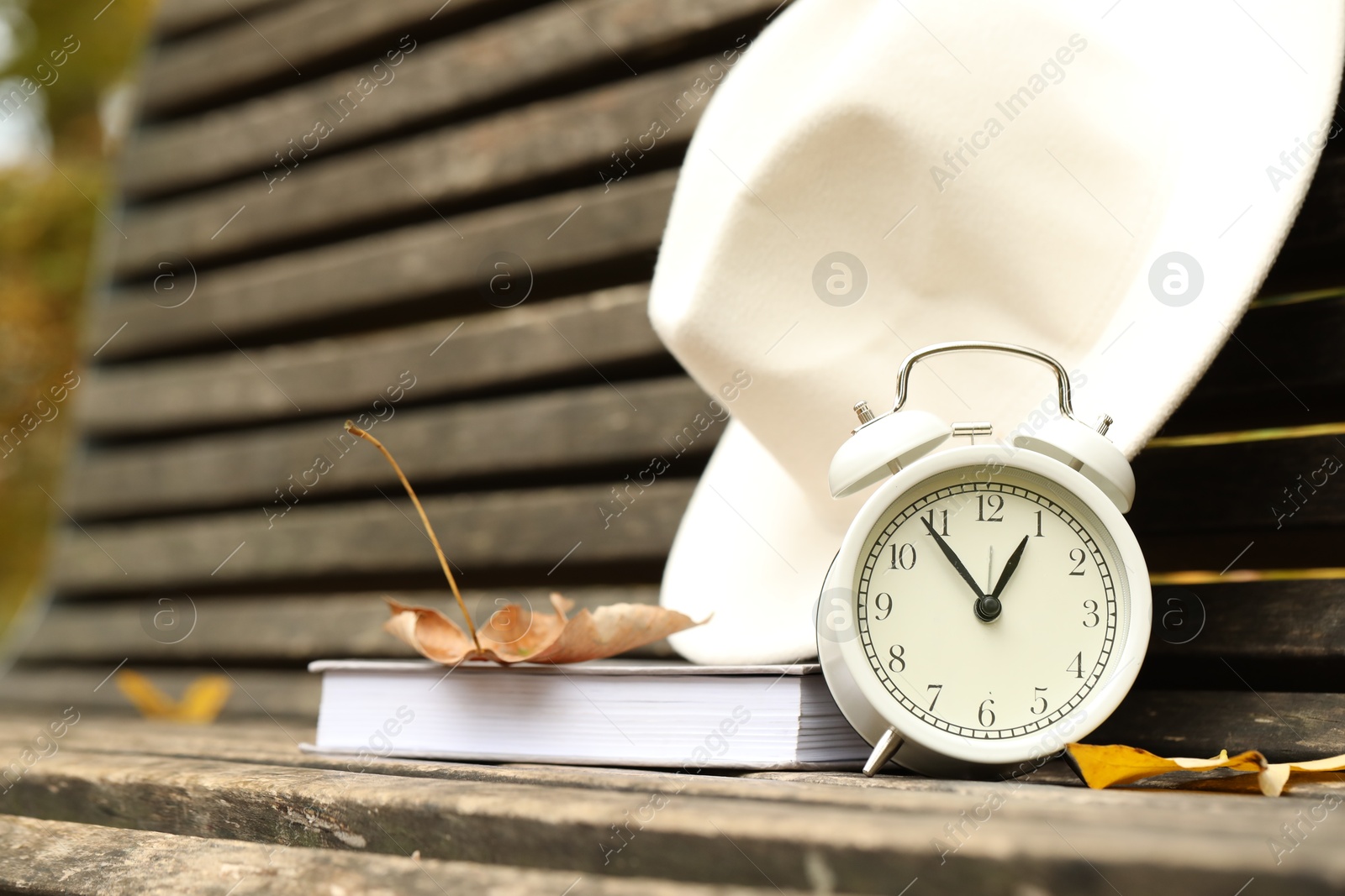 Photo of Autumn time. Alarm clock, hat and book on bench in park, closeup