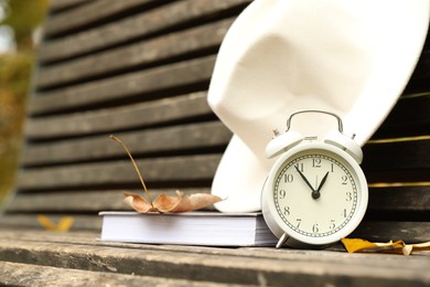 Photo of Autumn time. Alarm clock, hat and book on bench in park, closeup