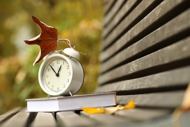 Photo of Autumn time. Alarm clock and book on bench in park, closeup