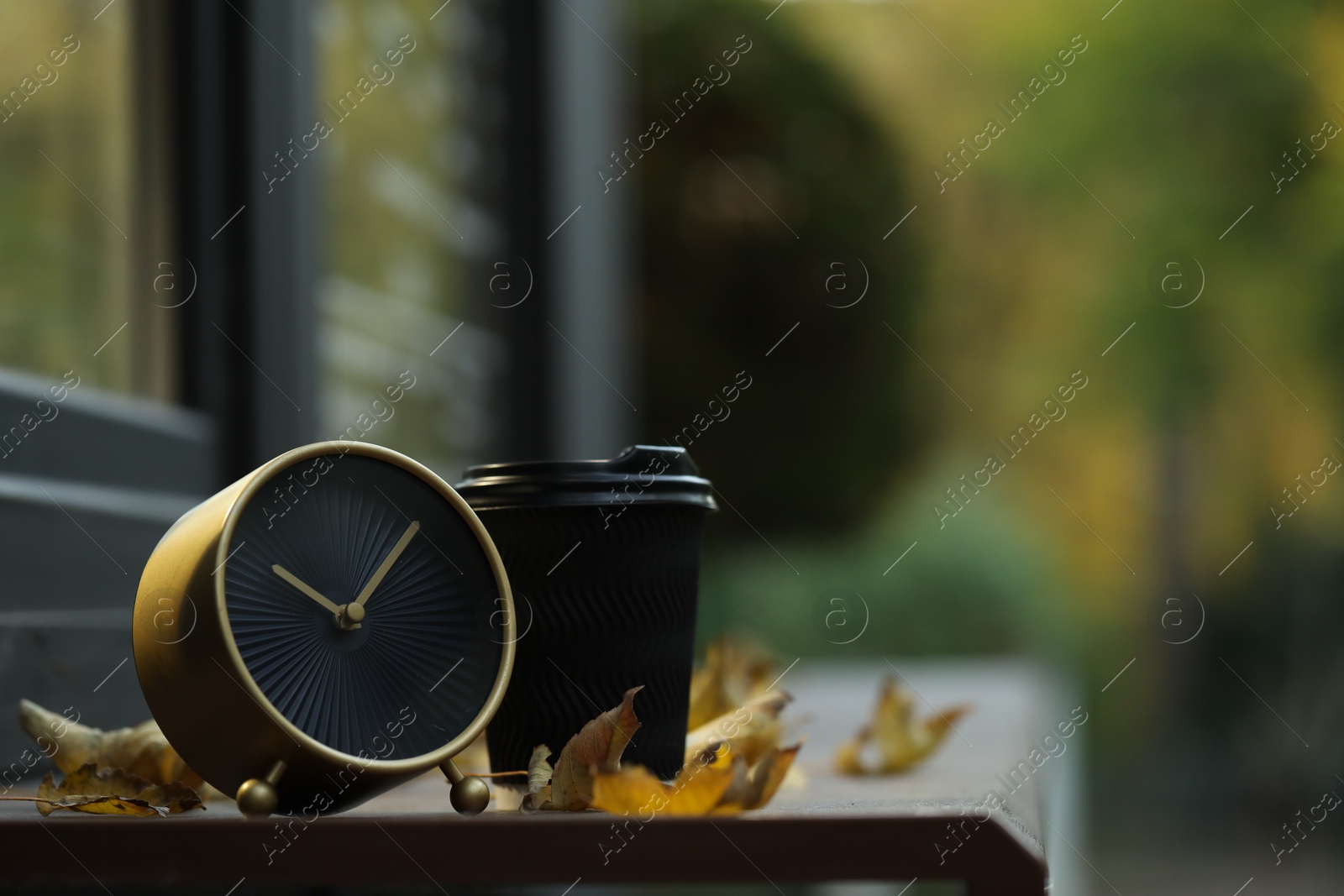 Photo of Autumn time. Alarm clock, fallen leaves and cup with hot drink near cafe outdoors, space for text