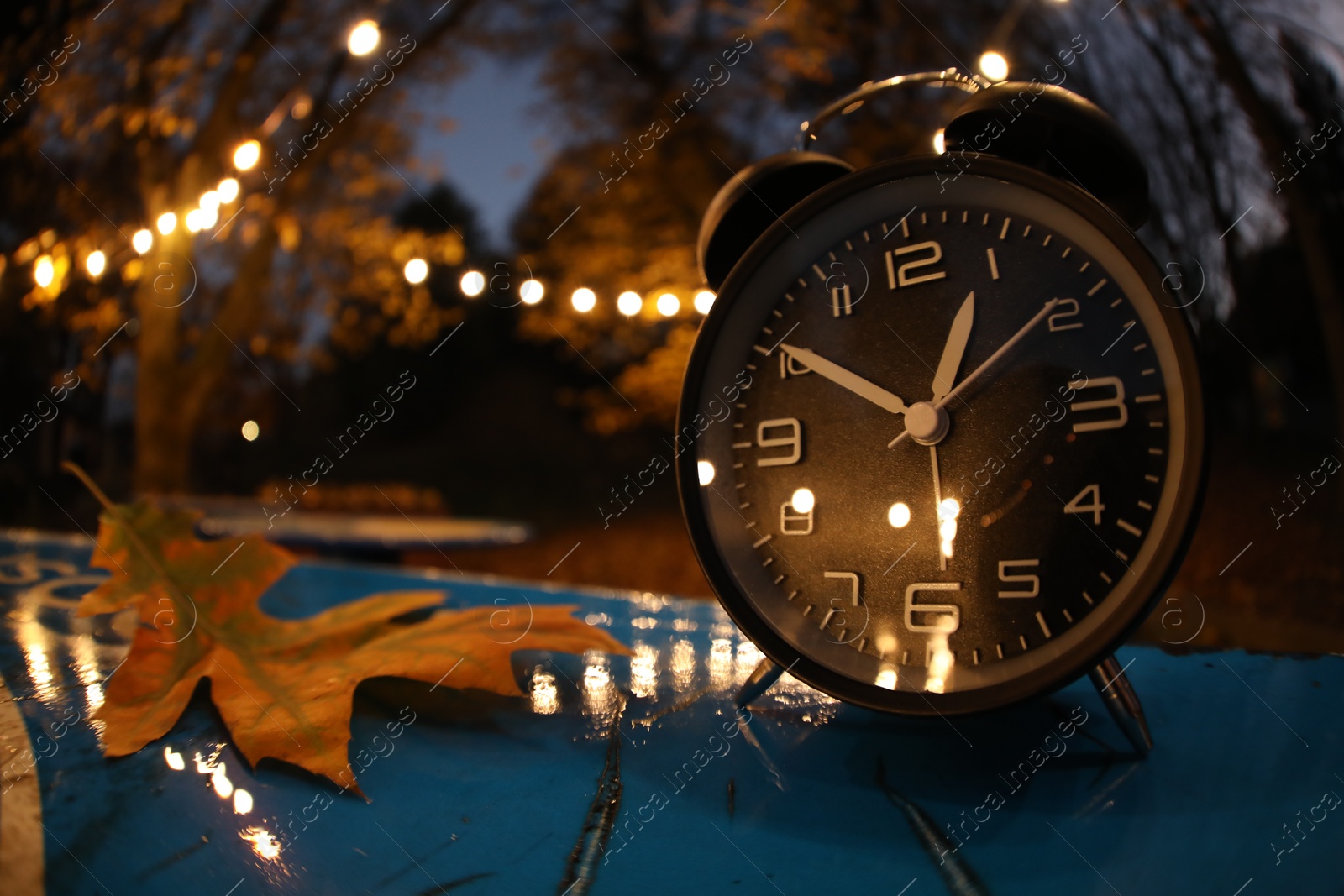 Photo of Autumn time. Alarm clock and fallen leaves on blue surface in evening park, wide angle lens