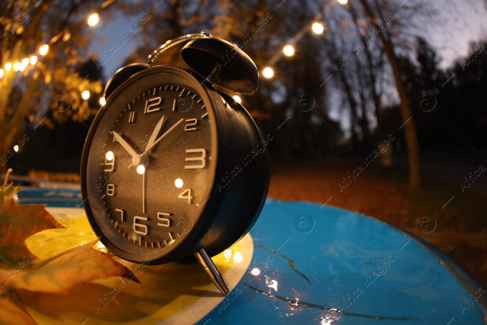 Photo of Autumn time. Alarm clock and fallen leaves on blue surface in evening park, wide angle lens