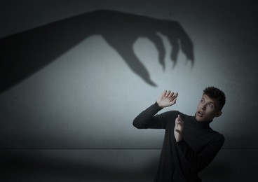 Image of Young man scared by monstrous hand reaching for him. Creepy shadow on grey wall