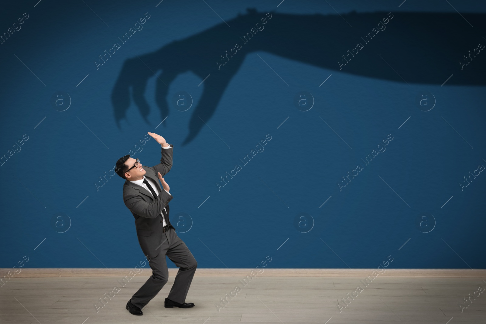 Image of Businessman scared by monstrous hand reaching for him. Creepy shadow on blue wall