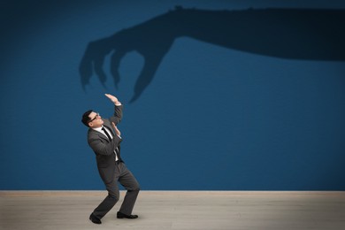 Image of Businessman scared by monstrous hand reaching for him. Creepy shadow on blue wall