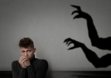 Young man scared by hands of monster reaching for him. Creepy shadow on grey wall