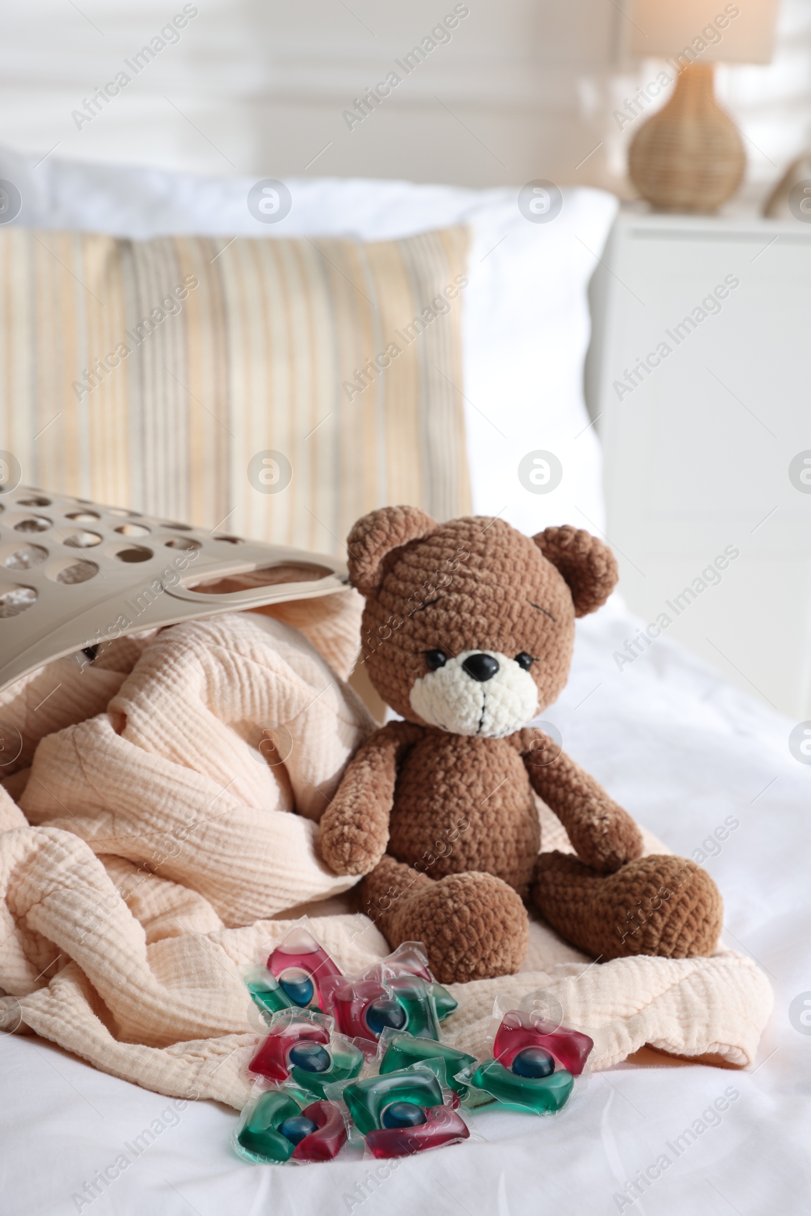 Photo of Laundry detergent capsules, teddy bear and basket with clean linens on bed indoors