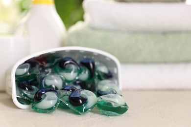 Photo of Container with laundry capsules, detergents and clean towels on table against blurred background, selective focus