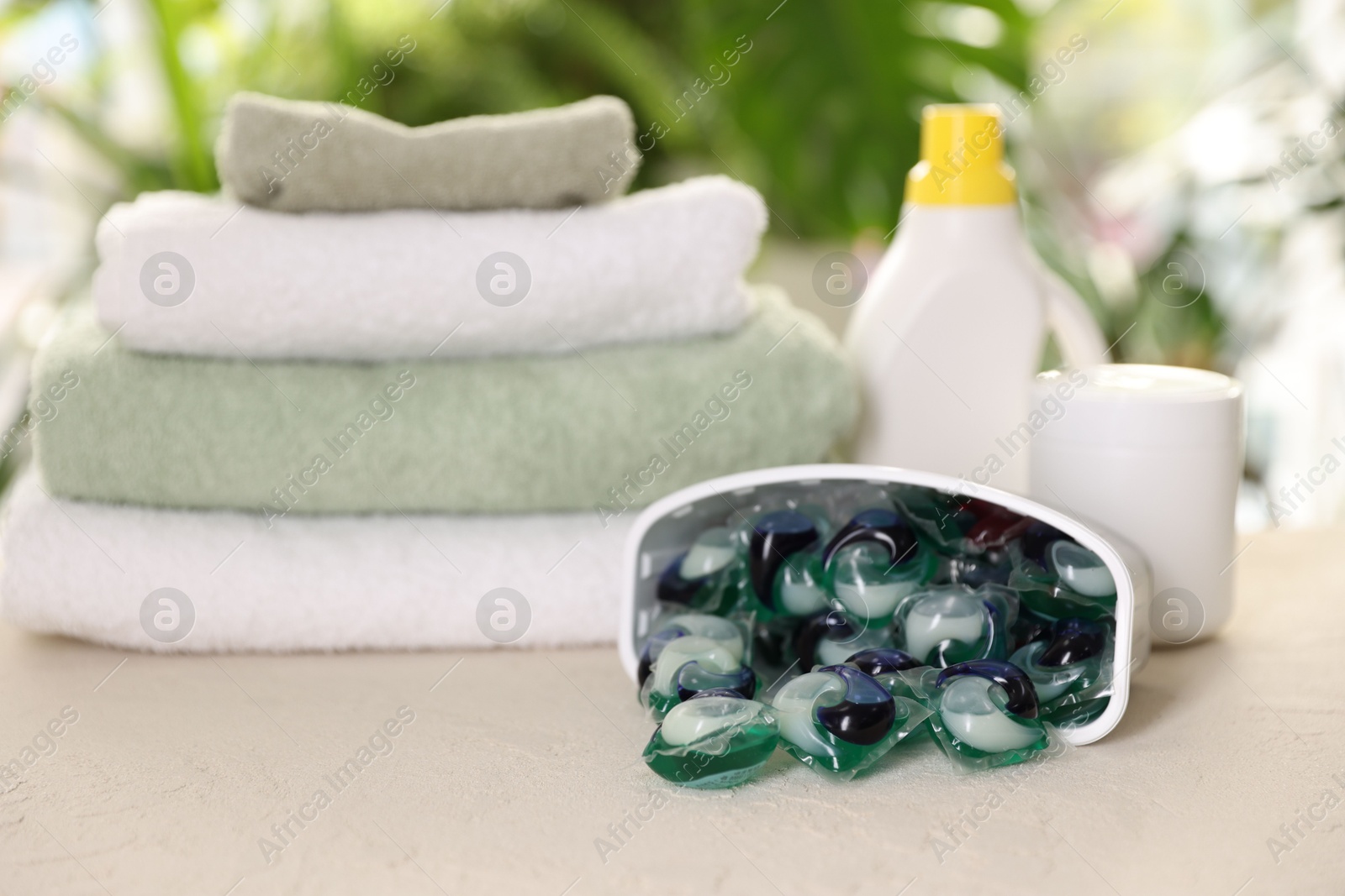 Photo of Container with laundry capsules, detergents and clean towels on table against blurred background, selective focus