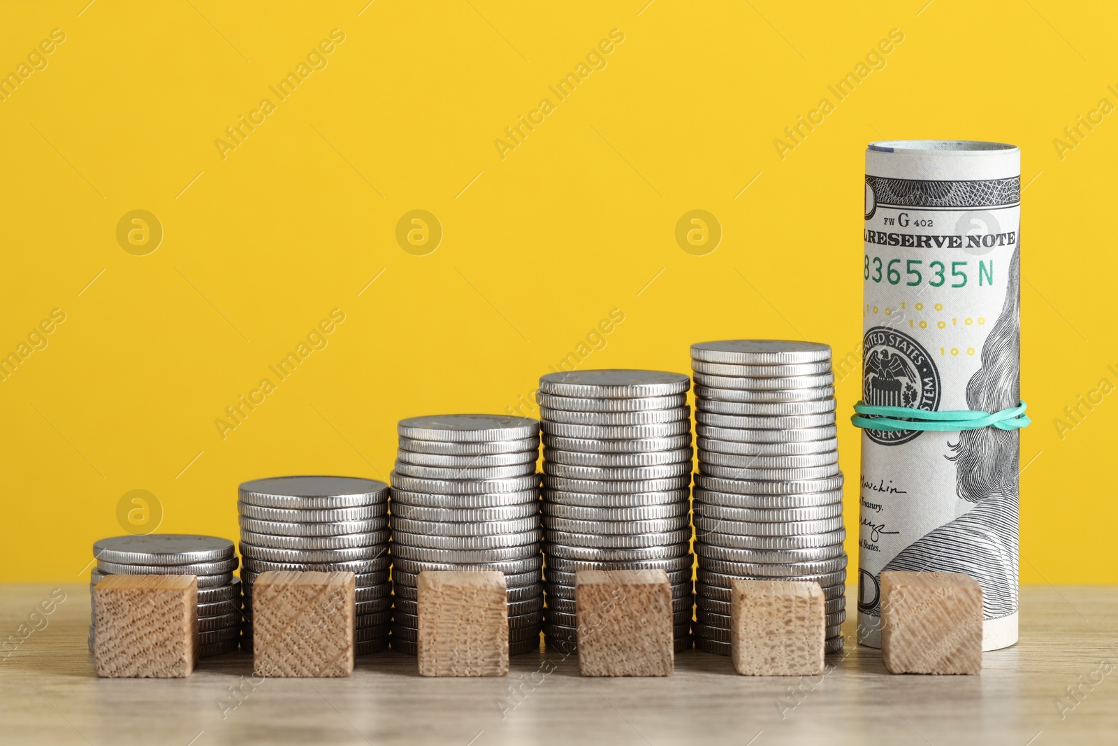 Photo of Blank cubes, stacked coins and dollar banknotes on wooden table against yellow background, closeup. Salary concept