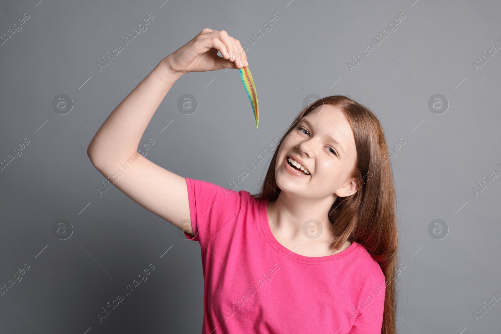 Photo of Happy teenage girl with tasty rainbow sour belt on grey background