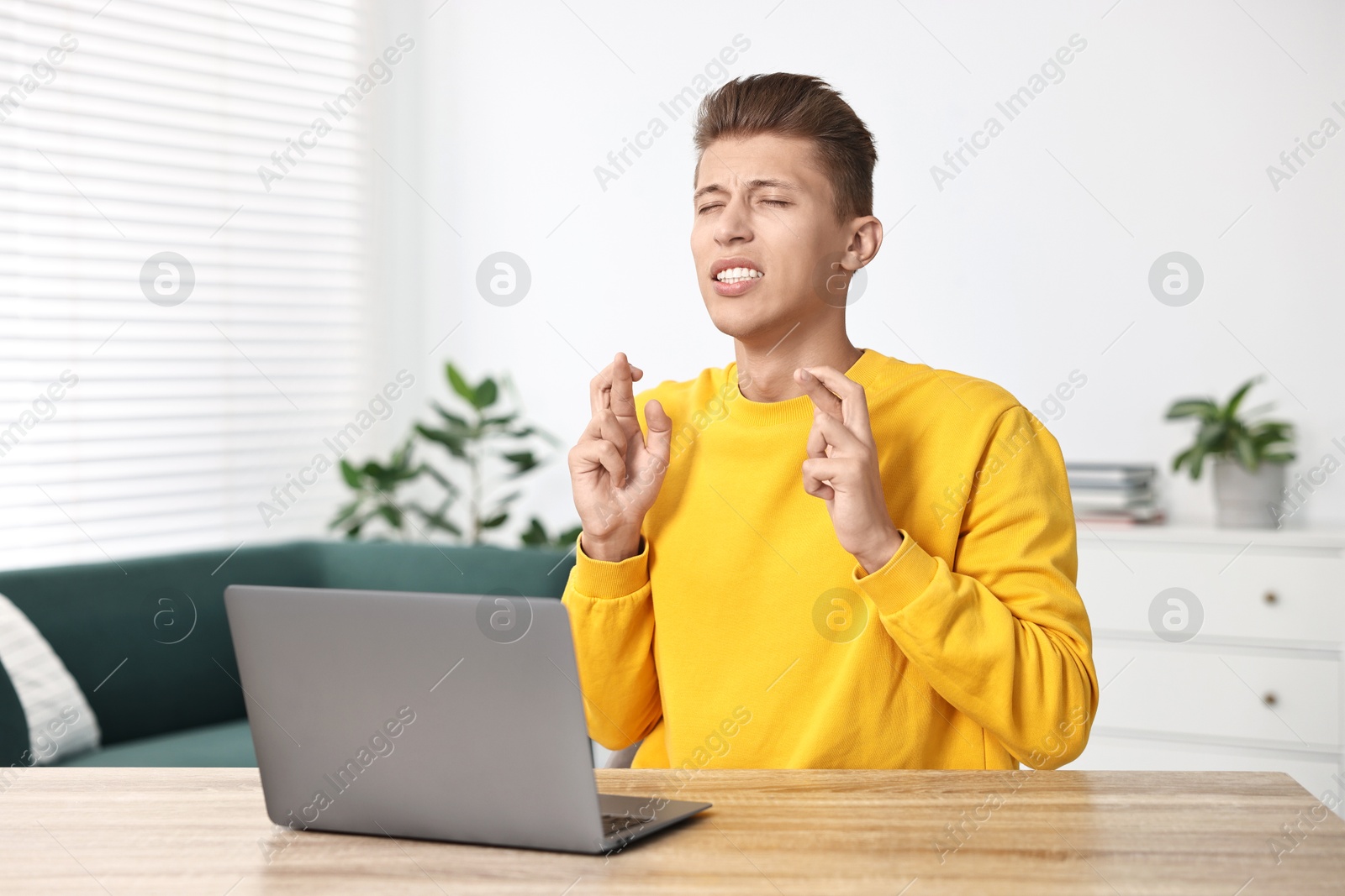 Photo of Worried student with crossed fingers at table with laptop indoors. Hope for good exam result