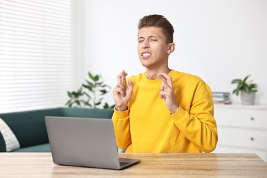 Photo of Worried student with crossed fingers at table with laptop indoors. Hope for good exam result