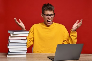 Photo of Emotional student having stress before exam with stack of books and laptop at wooden table against red background