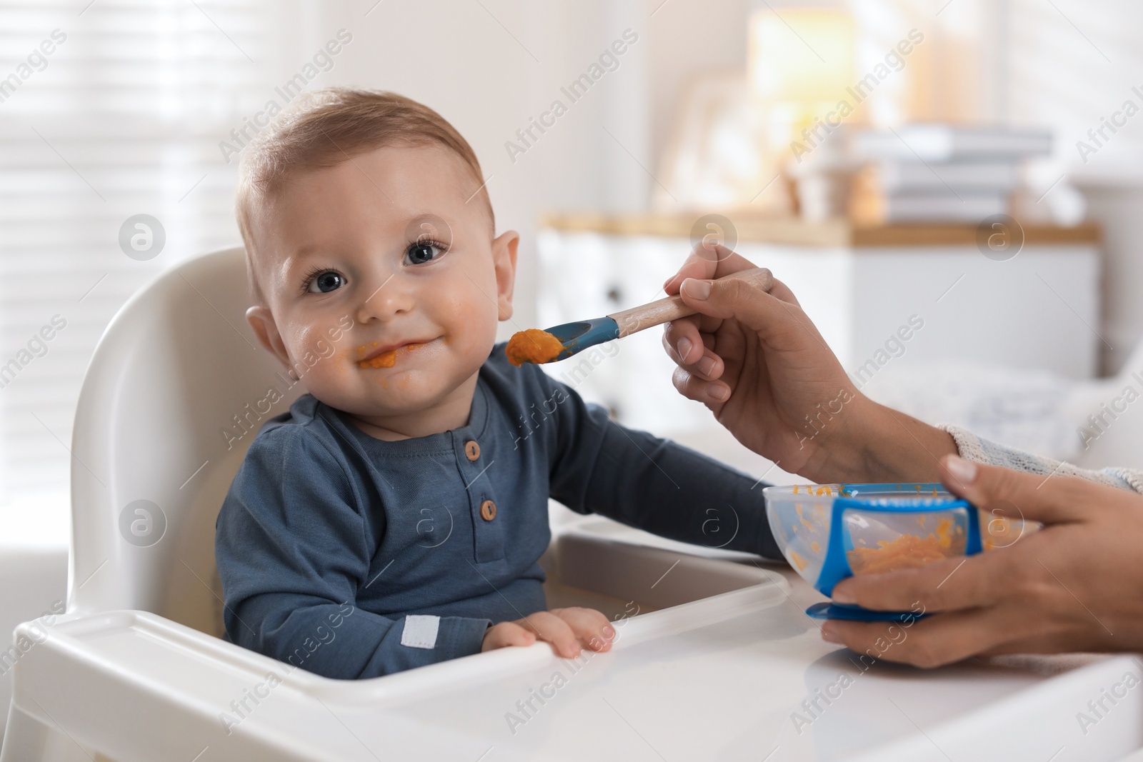 Photo of Mother feeding her cute little baby in high chair at home, closeup