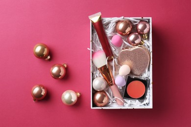 Photo of Christmas present. Decorative cosmetics in gift box and festive balls on dark pink background, top view