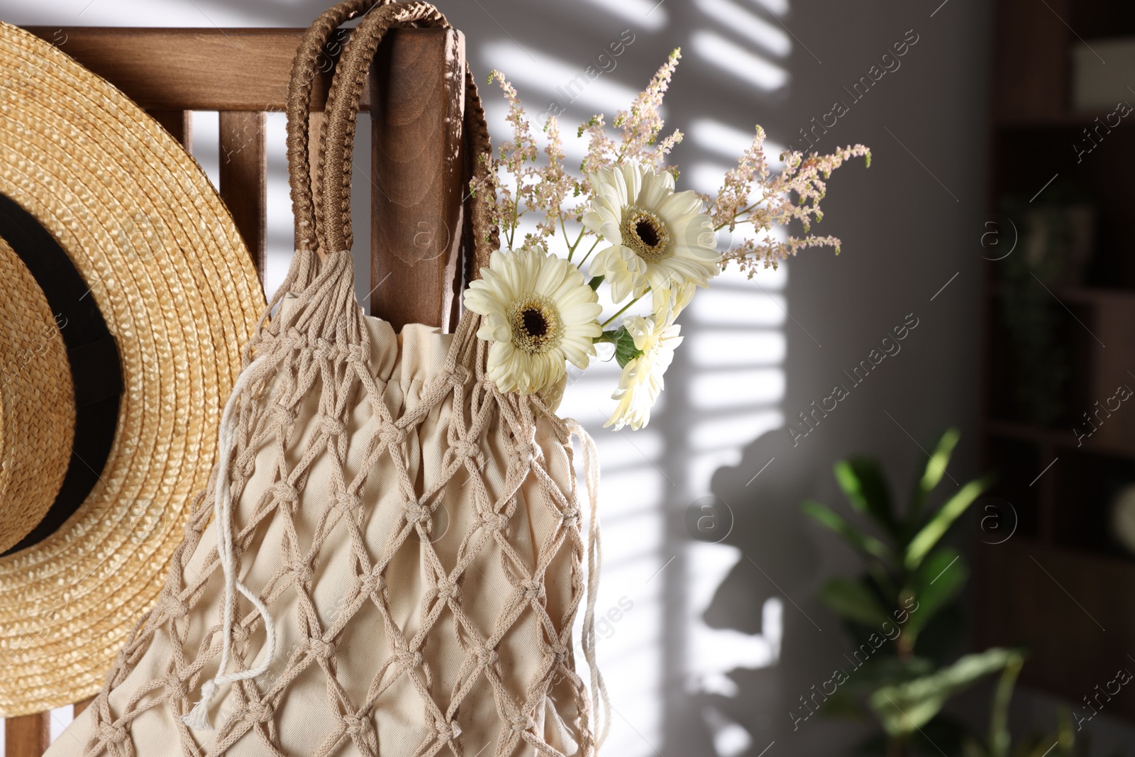 Photo of Handmade macrame shopping bag with flowers and straw hat on wooden chair indoors, space for text