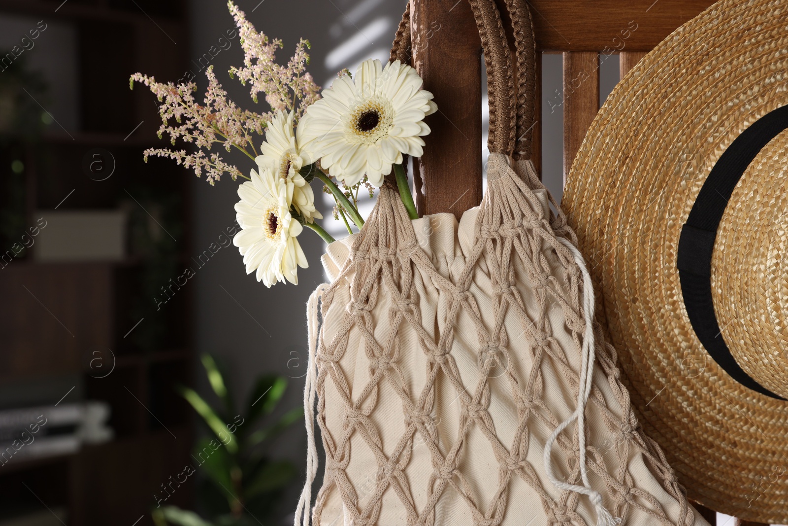 Photo of Handmade macrame shopping bag with flowers and straw hat on wooden chair indoors