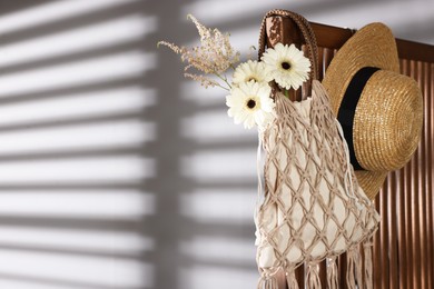 Photo of Handmade macrame shopping bag with flowers and straw hat on wooden chair indoors, space for text